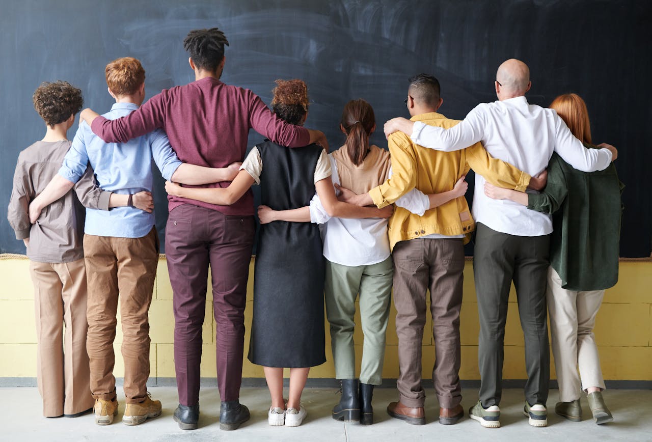 hero-img A diverse group of adults in casual outfits hugging in front of a chalkboard, symbolizing teamwork.
